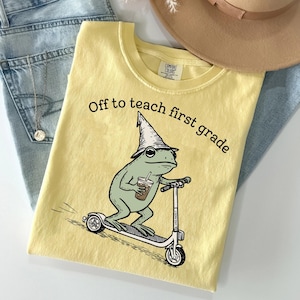 May include: A pale yellow t-shirt featuring a graphic of a green frog wearing a pointed hat, holding a coffee cup, and riding a scooter. The text above the frog reads, "Off to teach first grade." The shirt is displayed with blue jeans and a hat.