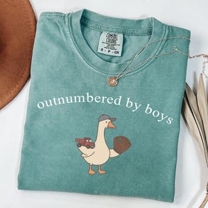 May include: Teal Comfort Colors t-shirt with the phrase "outnumbered by boys" in white. The shirt features a cartoon goose wearing a baseball cap and holding a baseball glove and a red truck. A silver necklace with a small pendant is on the shirt.