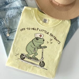May include: A pale yellow t-shirt featuring a graphic of a green frog wearing a nurse cap and a stethoscope while riding a scooter. The text reads OFF TO HELP LITTLE PATIENTS with three small hearts. The shirt has a Comfort Colors brand label.