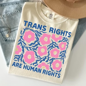 Könnte beinhalten: Cremefarbenes T-Shirt mit dem Text "TRANS RIGHTS ARE HUMAN RIGHTS" in Blau. Das Shirt hat ein Blumendesign mit rosa Blüten und blauen Blättern. Das Shirt liegt gefaltet auf einer blauen Jeans.