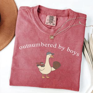 May include: A dusty rose t-shirt with the phrase "outnumbered by boys" in white. The shirt features a cartoon goose wearing a baseball cap and glove, holding a toy truck. A gold necklace is draped across the shirt.