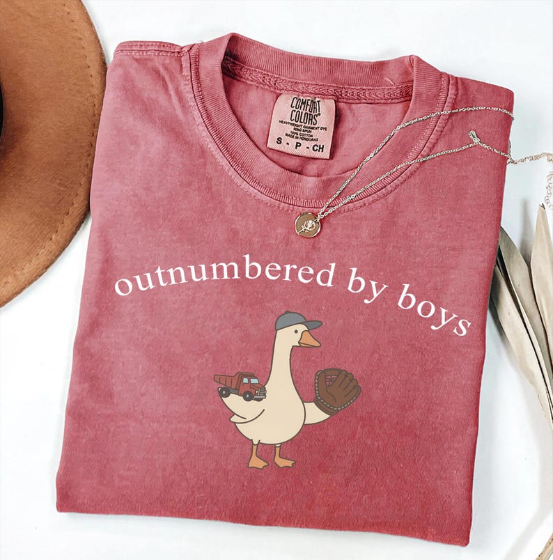 May include: A dusty rose t-shirt with the phrase "outnumbered by boys" in white. The shirt features a cartoon goose wearing a baseball cap and glove, holding a toy truck. A gold necklace is draped across the shirt.