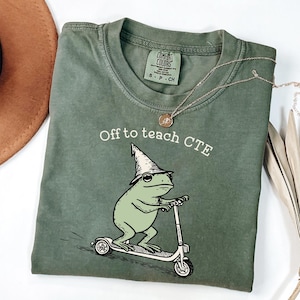 Könnte beinhalten: Olivgrünes T-Shirt mit dem Text "Off to teach CTE" und einem Cartoon-Frosch auf einem Roller, der einen Zaubererhut trägt. Eine goldene Halskette liegt über dem Shirt.