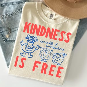 Puede incluir: Camiseta color crema con las palabras rojas "KINDNESS IS FREE" e ilustraciones azules. La camiseta también tiene la frase "sprinkle it everywhere". Un sombrero beige y unos vaqueros azules están en el fondo.