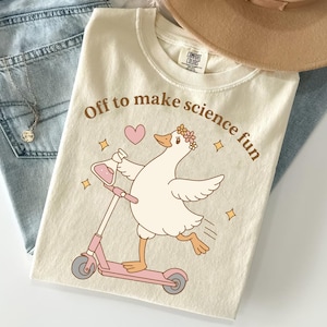 May include: A cream-colored t-shirt featuring a graphic of a white goose wearing a flower crown, riding a pink scooter, and holding a pink liquid-filled flask. The text "Off to make science fun" is printed in brown above the goose with a pink heart.