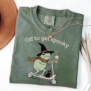 May include: An olive green t-shirt featuring a graphic of a frog wearing a black witch hat and a plaid scarf, riding a scooter while holding a coffee cup labeled PSL. A small black cat sits on the scooter. The text reads Off to get spooky.
