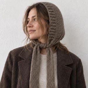Könnte beinhalten: Eine braune Strickhaube mit geripptem Rand und langen Bändern, die unter dem Kinn geknotet werden. Die Kopfbedeckung besteht aus strukturiertem Garn und wird über einem hellen Pullover und einem dunkelbraunen Wollmantel mit Reverskragen getragen.
