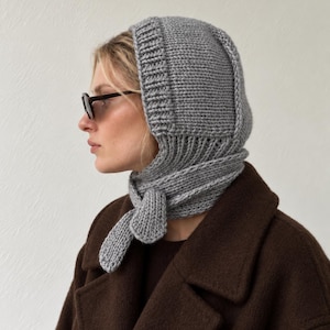 Könnte beinhalten: Eine graue Strickhaube mit gerippter Textur und einer Bindung am Hals. Die Kopfbedeckung zeichnet sich durch ein dickes, strukturiertes Strickmuster aus, das den Kopf bedeckt und um das Kinn reicht, befestigt durch zwei Strickbänder.