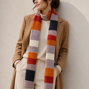 Könnte beinhalten: Ein langer Strickschal mit horizontalen Streifen in abwechselnden Farben: Dunkelmarineblau, leuchtendes Orange, kräftiges Rot, sanftes Creme und gedämpftes Lavendel. Der Schal liegt um den Hals und hängt in zwei langen, gleichmäßigen Teilen herab.