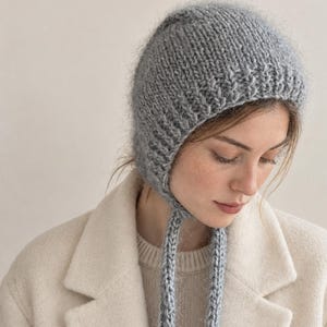 Könnte beinhalten: Eine hellgraue Strickhaube mit einer flauschigen, strukturierten Oberfläche und langen, herabhängenden geflochtenen Bändern. Die Kopfbedeckung hat ein Rippenmuster am unteren Rand und besteht aus einem weichen Material mit sichtbarem Strickmuster.