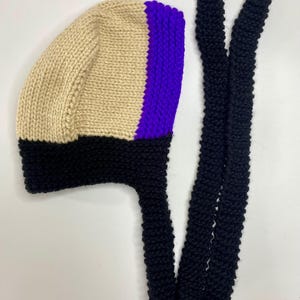 May include: A hand-knitted bonnet featuring a color-blocked design with a beige main section, a vibrant purple vertical stripe, and a thick black brim. The bonnet includes two long, black knitted ties extending from the base for secure fastening.