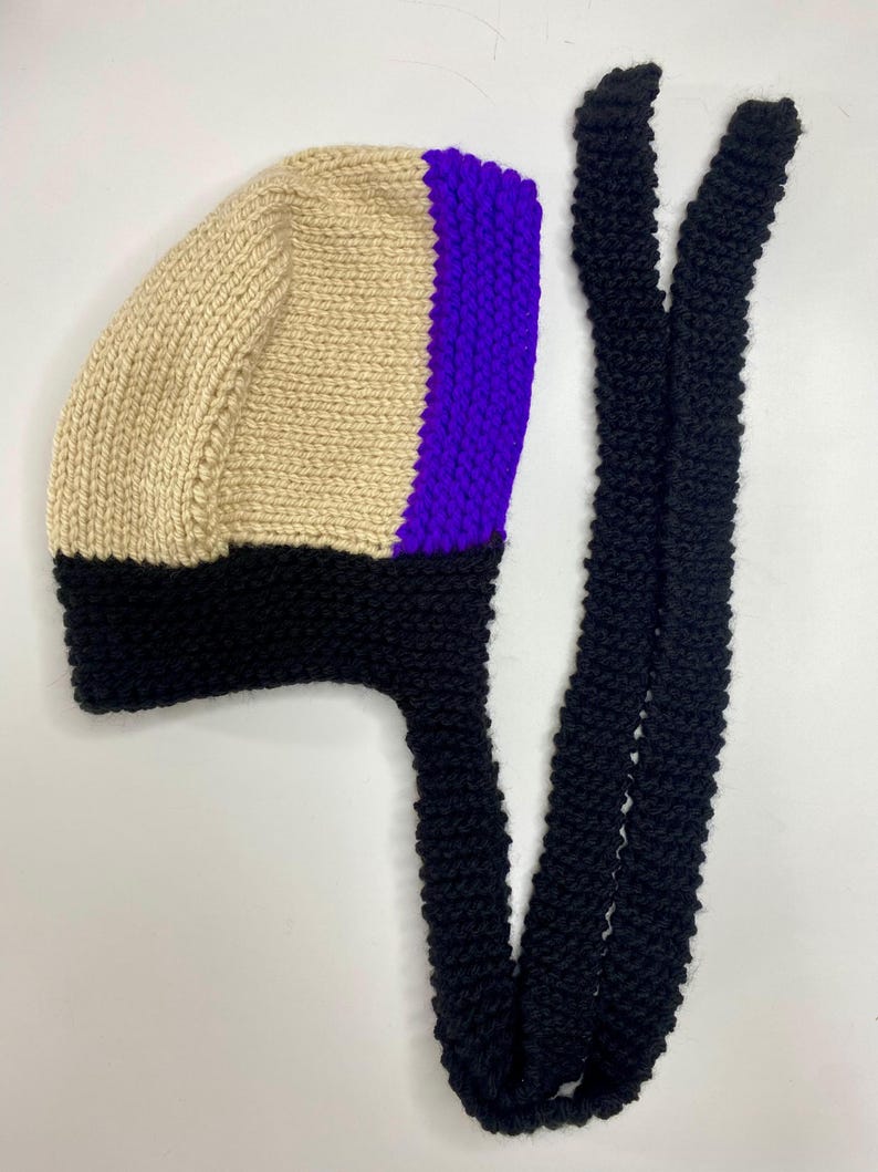 May include: A hand-knitted bonnet featuring a color-blocked design with a beige main section, a vibrant purple vertical stripe, and a thick black brim. The bonnet includes two long, black knitted ties extending from the base for secure fastening.