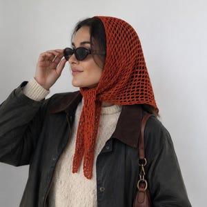Puede incluir: Un pañuelo de crochet color óxido con un patrón de red de diamantes, usado sobre un suéter de punto color crema y una chaqueta verde oliva oscuro con cuello marrón. El conjunto incluye gafas de sol ovaladas oscuras y un bolso de hombro de cuero marrón.