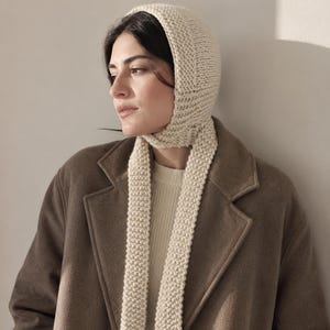 May include: A cream-colored knitted bonnet with long, dangling ties that drape down the front. The headpiece features a textured garter stitch pattern, providing a snug fit around the head and neck, worn over a ribbed cream sweater and a brown coat.