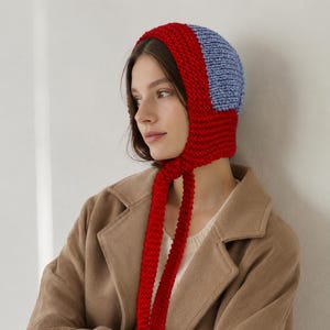 May include: A chunky knit bonnet featuring a vibrant red border and a light blue center panel. The headwear is constructed with a thick, textured yarn and includes two long, matching red knitted ties that hang down from the chin area.