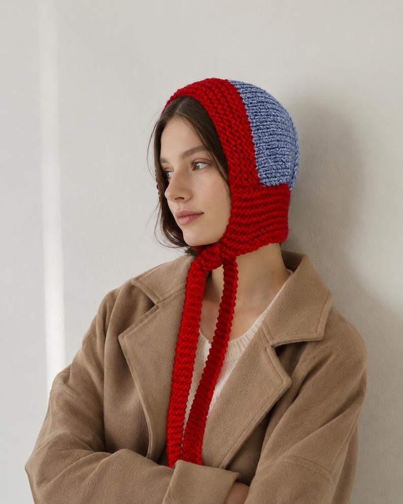 May include: A chunky knit bonnet featuring a vibrant red border and a light blue center panel. The headwear is constructed with a thick, textured yarn and includes two long, matching red knitted ties that hang down from the chin area.