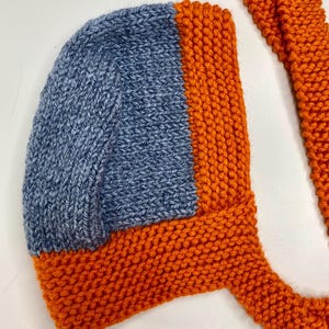 May include: A hand-knitted bonnet featuring a blue stockinette stitch center panel framed by a vibrant orange garter stitch border and long matching orange ties. The headwear is constructed with a structured shape and textured yarn finish.