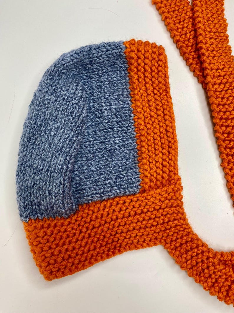 May include: A hand-knitted bonnet featuring a blue stockinette stitch center panel framed by a vibrant orange garter stitch border and long matching orange ties. The headwear is constructed with a structured shape and textured yarn finish.