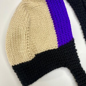 May include: A hand-knitted bonnet featuring a color-blocked design with a large cream-colored section, a vibrant purple vertical stripe, and a solid black border along the bottom edge, complete with long black knitted ties extending from the sides.