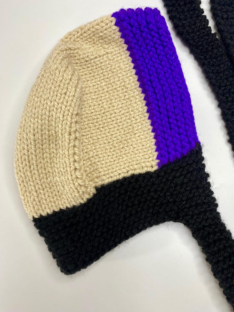 May include: A hand-knitted bonnet featuring a color-blocked design with a large cream-colored section, a vibrant purple vertical stripe, and a solid black border along the bottom edge, complete with long black knitted ties extending from the sides.