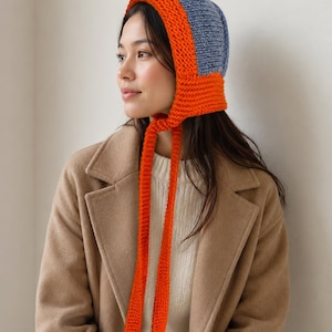 May include: A knit bonnet featuring a two-tone design with a bright orange border and ties, contrasted by a textured blue crown. The bonnet is constructed with a thick, chunky yarn knit pattern and includes long, dangling orange ties at the chin.