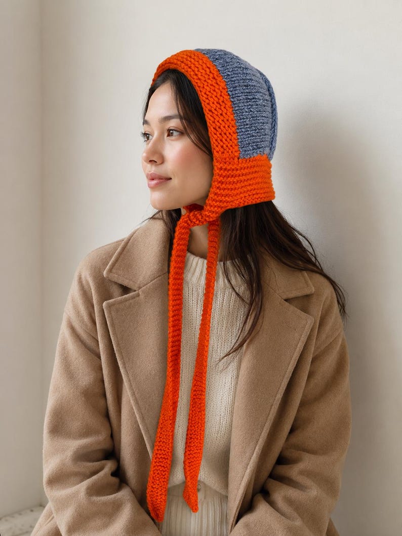 May include: A knit bonnet featuring a two-tone design with a bright orange border and ties, contrasted by a textured blue crown. The bonnet is constructed with a thick, chunky yarn knit pattern and includes long, dangling orange ties at the chin.