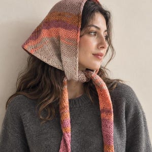 Puede incluir: Un pañuelo tejido a mano con una paleta de colores variados en tonos rosa suave, naranja y beige. El accesorio de forma triangular está confeccionado con un punto de tejido texturizado e incluye dos lazos largos integrados que cuelgan.