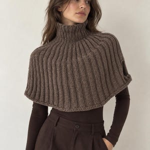 Könnte beinhalten: Ein brauner Strickponcho mit einem hohen, gerippten Rollkragen und einer durchgehenden vertikalen Rippenstruktur. Das Kleidungsstück ist kurz geschnitten, endet oberhalb der Taille und verfügt über seitliche Öffnungen für die Arme.