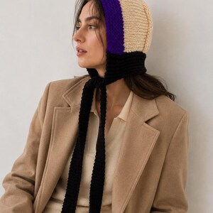May include: A knit bonnet featuring a color-blocked design with a cream-colored crown, a vibrant purple band framing the face, and a black base with long, dangling black knit ties that hang down from the chin area to secure the head covering.