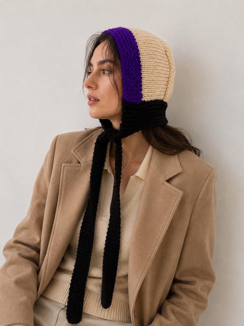 May include: A knit bonnet featuring a color-blocked design with a cream-colored crown, a vibrant purple band framing the face, and a black base with long, dangling black knit ties that hang down from the chin area to secure the head covering.