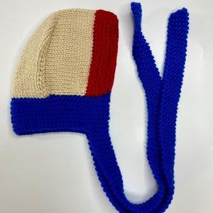 May include: A hand-knitted bonnet featuring a color-blocked design with a cream-colored main section, a vertical red stripe on the side, and a vibrant blue border along the bottom edge. The bonnet includes two long, matching blue knitted ties.