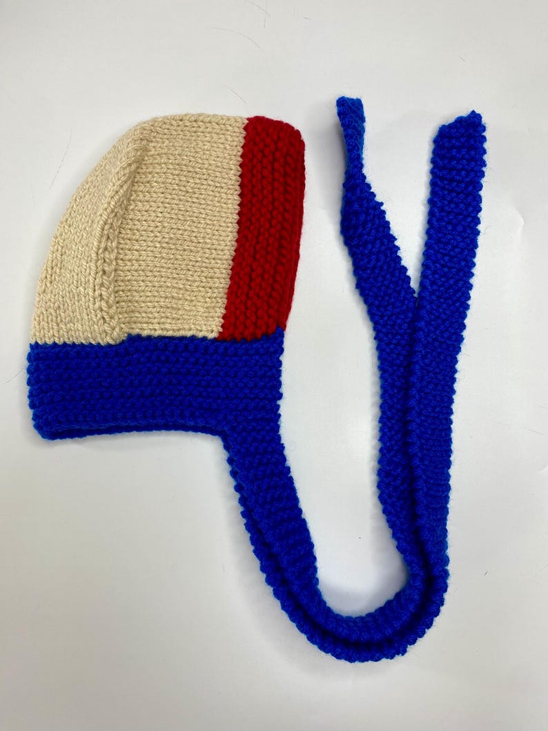 May include: A hand-knitted bonnet featuring a color-blocked design with a cream-colored main section, a vertical red stripe on the side, and a vibrant blue border along the bottom edge. The bonnet includes two long, matching blue knitted ties.