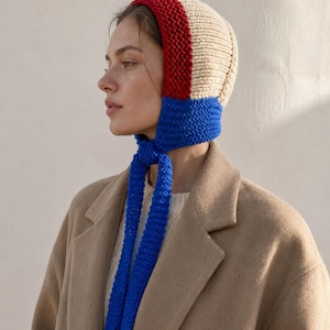 May include: A knitted bonnet featuring a colour-blocked design with a cream-coloured crown, a red band along the front edge, and a bright blue chin strap that ties under the chin. The headwear is crafted with a textured, chunky knit stitch pattern.