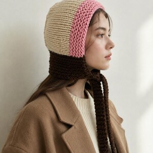 May include: A color-blocked knit bonnet featuring a cream-colored crown, a pink band around the face, and a dark brown neck section with long, thick knitted ties that hang down. The bonnet is constructed with a visible, chunky yarn texture.