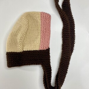 May include: A hand-knitted bonnet featuring a color-blocked design with a cream-colored crown, a soft pink vertical stripe, and a dark brown base and trim. The bonnet includes long, matching dark brown knitted ties for securing under the chin.