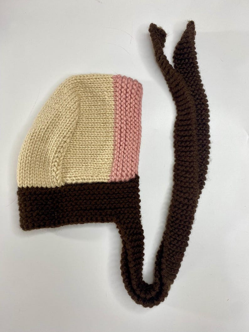 May include: A hand-knitted bonnet featuring a color-blocked design with a cream-colored crown, a soft pink vertical stripe, and a dark brown base and trim. The bonnet includes long, matching dark brown knitted ties for securing under the chin.