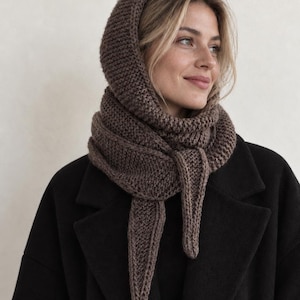 Könnte beinhalten: Ein dunkelbrauner Strickkapuzenschal mit einem dicken, strukturierten Kraus-Rechts-Muster. Das Accessoire umschließt Hals und Kopf und hat zwei lange, spitz zulaufende Enden, die über einem schwarzen Mantel mit breitem Revers nach vorne hängen.