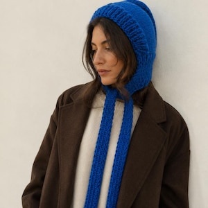 May include: A bright blue knitted bonnet with a ribbed texture and two long, matching knitted ties hanging down from the sides. The bonnet features a thick, structured knit pattern that covers the head and ears, providing a bold pop of color.