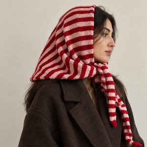 May include: A knitted headscarf featuring horizontal stripes in alternating shades of red and light pink. The fabric has a visible ribbed texture and is tied securely under the chin, draping over the head and shoulders in a triangular shape.