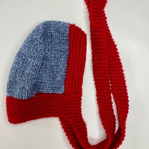 May include: A hand-knitted bonnet featuring a light blue textured crown and a vibrant red border. The bonnet includes two long, matching red knitted ties extending from the sides, designed to be fastened under the chin for a secure fit.