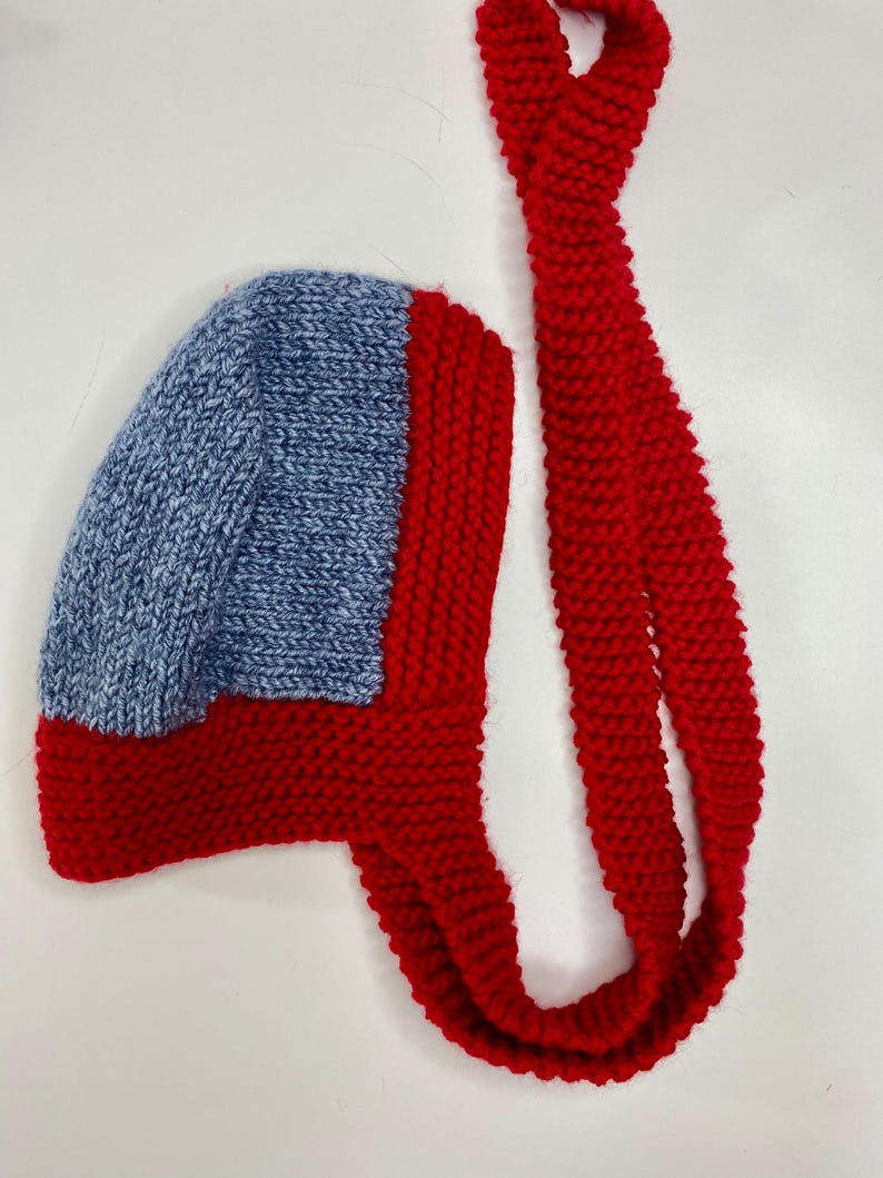 May include: A hand-knitted bonnet featuring a light blue textured crown and a vibrant red border. The bonnet includes two long, matching red knitted ties extending from the sides, designed to be fastened under the chin for a secure fit.