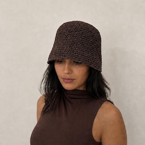 Pode incluir: Um chapéu bucket castanho-escuro com uma estrutura de croché tecido e texturizado, apresentando uma copa arredondada e uma aba curta inclinada para baixo. O chapéu é feito de um material de fibra denso e uniforme com um acabamento mate.