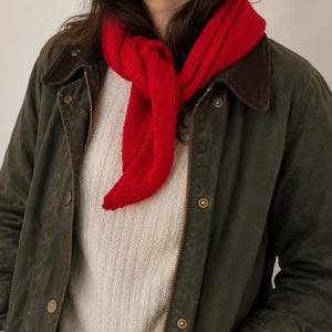 Könnte beinhalten: Ein leuchtend roter Strickschal, der einfach um den Hals geknotet ist, getragen über einem cremefarbenen Rippenstrickpullover und einer olivgrünen gewachsten Baumwolljacke mit dunkelbraunem Cordkragen und goldfarbenen Metalldruckknöpfen.