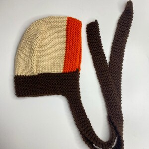 May include: A hand-knitted bonnet featuring a cream-colored main section, a vibrant orange vertical stripe, and a dark brown garter stitch border. The bonnet includes two long, matching dark brown knitted ties extending from the base for fastening.