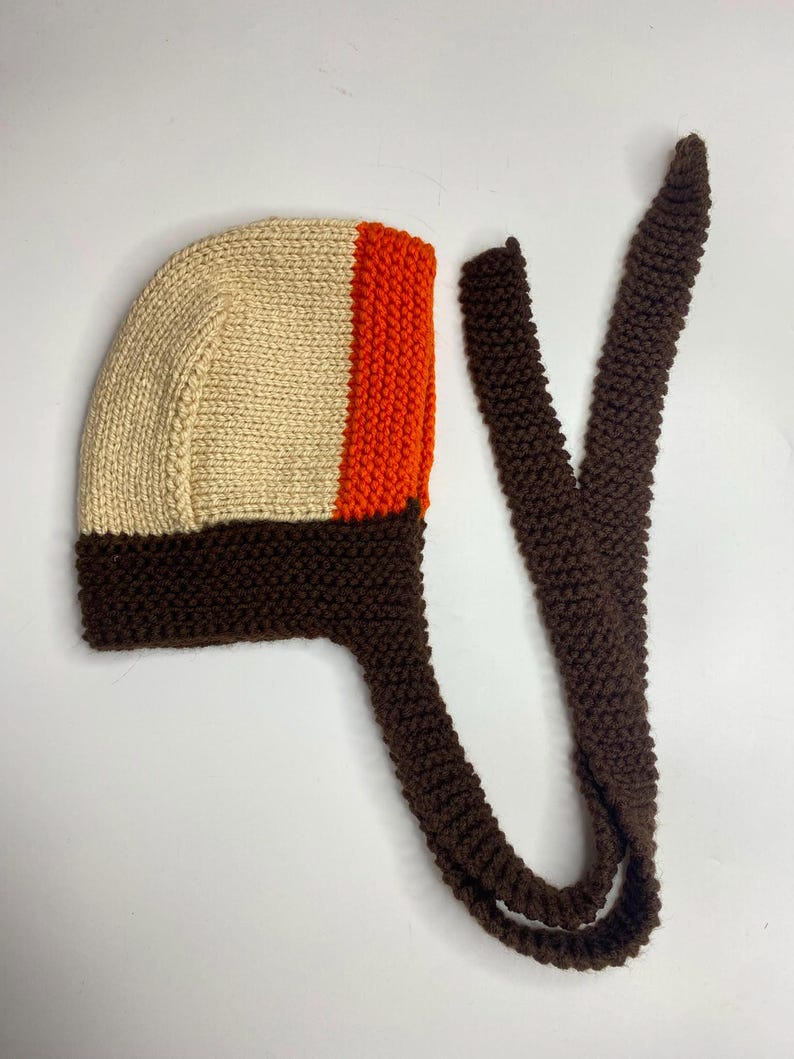 May include: A hand-knitted bonnet featuring a cream-colored main section, a vibrant orange vertical stripe, and a dark brown garter stitch border. The bonnet includes two long, matching dark brown knitted ties extending from the base for fastening.