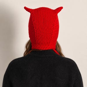 May include: A bright red hand-knitted beanie featuring two small, pointed horn-like protrusions on the top. The hat is constructed with a thick, textured yarn knit pattern and includes long, dangling knitted ties extending from the bottom sides.