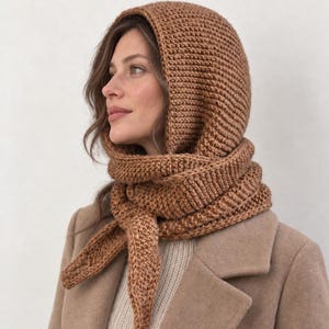Könnte beinhalten: Ein mittelbrauner Strick-Kapuzenschal mit einem strukturierten Rippenstrickmuster. Das Accessoire umschließt Kopf und Hals und wird mit langen Enden vorne verknotet, wodurch Kopf und Halsbereich vollständig bedeckt und geschützt werden.