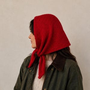 May include: A bright red knitted headscarf tied under the chin, featuring a ribbed texture. The headscarf is worn over a cream-coloured jumper and a dark olive green jacket with a brown corduroy collar, visible from the chest up.