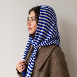Könnte beinhalten: Ein gestricktes Kopftuch mit einem Muster aus horizontalen königsblauen und cremefarbenen Streifen. Der Stoff ist über den Kopf drapiert und unter dem Kinn gebunden, mit zwei langen herabhängenden Enden, getragen über einem braunen Mantel.