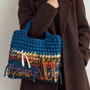 Può includere: Una borsa tote all'uncinetto fatta a mano con una sezione superiore blu profondo e una sezione inferiore multicolore con strisce di tessuto pendenti. La borsa è realizzata in filato spesso nei toni del blu, giallo, rosso, verde e marrone.